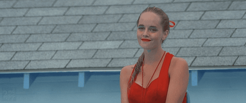 Movie gif. Marley Shelton as Wendy Peffercorn the Lifeguard in Sandlot sits on the lifeguard seat shaking her head with a warm smile on her face. She waves cutely to whoever she’s smiling at.