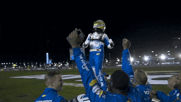 Sports gif. NASCAR driver Rick Stenhouse Jr. stands on the hood of a race car, pumping his hands in the air, then jumping down to body surf on his cheering teammates. The camera is orbiting around the scene, showing an empty track, stadium lights, and a stadium full of fans. 
