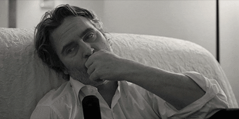 Movie gif. Joaquin Phoenix as Johnny in C'mon C'mon rests his head on a couch as he looks around sleepily while gnawing on something in his fingers.