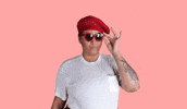 Video gif. Woman lowers her heart-shaped sunglasses, peering out with intrigue, blinking and smiling.