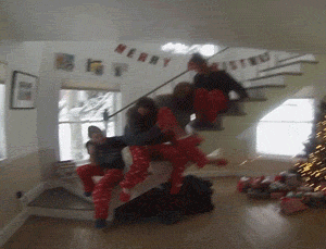 Video gif. Teenagers wearing Christmas pajamas sit on the edge of a staircase without a railing as two topple off in a room decorated for the holidays.