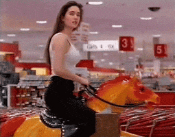 Video gif. A woman in a department store rides on a coin-operated pony ride, looking at us seductively.
