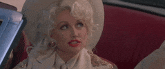 Movie gif. From The Best Little Whorehouse in Texas, Dolly Parton as Mona sits in the driver's seat of a convertible wearing a cowboy hat as she looks up and winks at someone with a growing smile. 