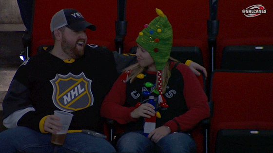 Video gif. Couple in the audience of a game look at each other giggling. The woman wearing a Christmas tree hat pretends to punch the man and he flinches, and laughs harder.