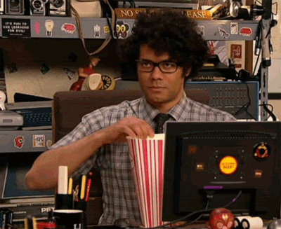 TV gif. Richard Ayoade as Maurice in The IT Crowd sits at his desk and looks smugly at someone off screen as he eats a piece of popcorn. 