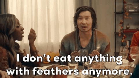 SNL gif. Simu Liu as a Thanksgiving dinner guest sits at a table holding silverware as he elaborates with his hands and says, "I don't eat anything with feathers anymore." Ego Nwodim as the dinner host cups a hand over her mouth in shock beside him.