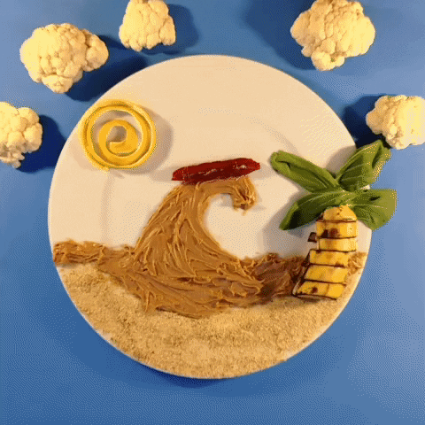 Stop motion gif. Plate of food depicting a tropical ocean scene, with a giant wave made of peanut butter, with a candy surfboard that a nutter butter drops onto briefly, as the lettuce-leaf palm fronds flutter and cauliflower clouds roll by.