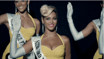 Music video gif. Beyonce in her video for Pretty Hurts. She's dressed as a beauty queen and she smiles gorgeously while waving one hand.