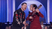 Academy of Country Music Awards gif. Noah Reid and Ashley McBryde performa duet where Reid sings into a standing mic with an expression as if he's reading off a tele-prompter while McBryde strums a large ukelele making differing looks on her face as if shes reacting to what Noah is singing.