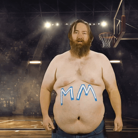 Video gif. Bearded man has the letters MM painted on his huge naked belly in front of a photo of a basketball court with a stadium full of fans, pumping his fist down like he's pumped. Text, "March Madness, Baby!'
