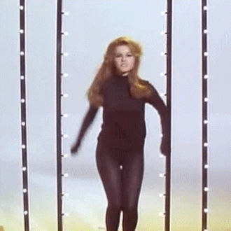 Video gif. A woman in all black dances, bringing her arms up and over her head to the beat. 