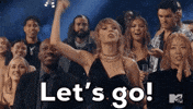 TV gif. Taylor Swift at the 2023 VMAs stands in the audience and pumps a finger high in the air, yelling "Let's go!" as other audience members look out in the same direction as Taylor as they clap and cheer. 