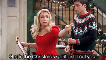 TV gif. Melissa Joan Hart from Melissa & Joey wears a red Christmas dress holds up a clipboard as she aggressively points and shouts at someone offscreen. Nick Robinson rolls up the sleeve of his sweater, maybe feeling a bit awkward. Text, “Get in the Christmas spirit or I’ll cut you!”