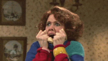 SNL gif. Kristen Wiig stands in a living room wearing a giant, multi-colored sweater which she pulls up over her mouth with both hands like she's trying to hide. Her mouth comes back out of it as she screams in horror. 