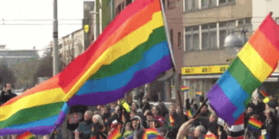Pride gif. Three large rainbow flags wave proudly in the air above a crowd waving smaller pride flags in the city street.
