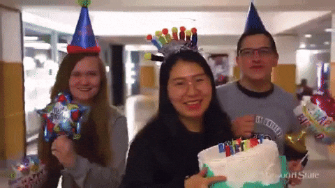 Video gif. Three students wearing Missouri State University shirts and birthday hats walk and dance towards us smiling, singing, and looking festive. One of them holds a birthday cake with candles on it and the two others hold balloons. 