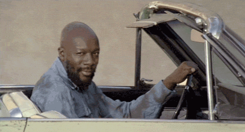 Movie gif. In a scene from Truck Turner, Isaac Hayes as Truck leans across the front passenger seat of his pale yellow convertible, smiles, and gives us a wiggling-fingers wave.
