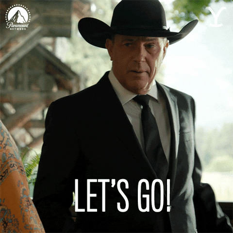 TV gif. Kevin Costner as John Dutton on Yellowstone wears a black cowboy hat and a black suit and tie. He turns toward someone, moving an object onto the counter next to him. With a serious expression on his face, he says, “Let’s go!”