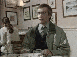 TV gif. Sitting at a restaurant table, Steve Coogan as Alan Partridge shrugs and frowns indifferently.