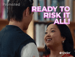 Sponsored gif. A smiling woman directs her excited gaze up towards a man as she speaks. They stand close together in conversation inside a bookstore next to a drawn animation of a heart with an arrow inside it.. Text reads, "Ready to risk it all!" with the Tinder logo in the bottom right corner.