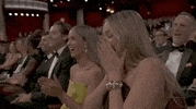 Oscars 2024 GIF. Margot Robbie, seated at the Oscars, covers her face with her hands, throwing her head back in laughter.