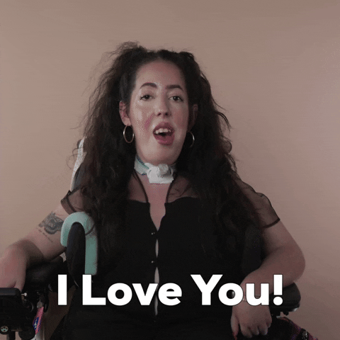Reaction gif. A Disabled white woman with muscular dystrophy with wavy brown half up half down with two pigtails on top, seated in her motorized wheelchair, shrugs her shoulders up to frame her face saying, "I love you."