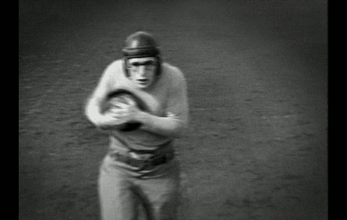 Movie gif. Carrying a football and wearing a leather football helmet, Harold Lloyd as Harold Lamb in The Freshman sprints towards us with a crouched neck.