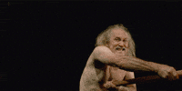 Video gif. Old man with long hair and wearing no shirt waves a stick that’s engulfed in flames. He grits his teeth angrily.