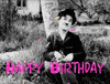 Movie gif. Black and white footage of Charlie Chaplin sitting on the grass against a tree tossing out his hands and bringing them back in with a shy or bashful expression. The video has been edited to make it look like he's throwing colorful confetti in the air. Text, "Happy Birthday."