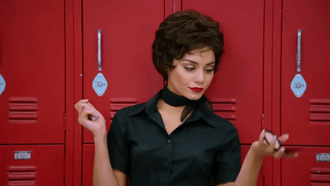 Movie gif. Vanessa Hudgens as Rizzo in Grease Live purses her lips, leaning against lockers, and looking up at herself in a compact mirror.