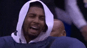 Sports gif. Kyrie Irving of the Cleveland Cavaliers sits on the sidelines with his hood up. He laughs hard and lowers his head to hide his laughter.