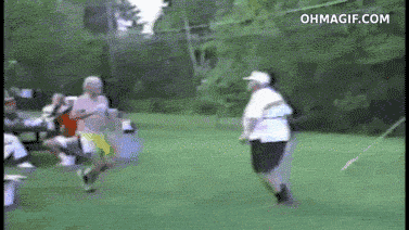 Video gif. At an outdoor gathering, a large-bellied man approaches another man and bumps bellies with him, which knocks the smaller guy over, who takes a few other people down with him.