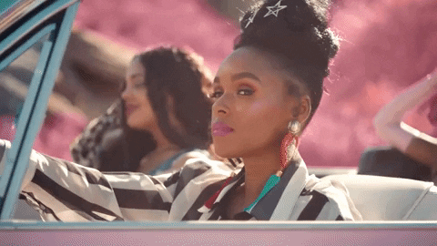 Music video gif. Janelle Monae in PYNK sits in the driver's seat of a pink convertible next to a woman in the passenger seat. She turns her head towards us calmly and looks into the distance. 