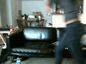 Video gif. Woman leaps onto a black leather couch and then tries to hop over the back, but falls on the ground, knocking stuff off a small table.