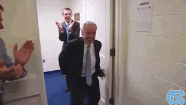 Sports gif. Roy Williams, head coach of the North Carolina Tarheels comes into the locker room dancing and the entire team gets hype as they all start jumping with one another.