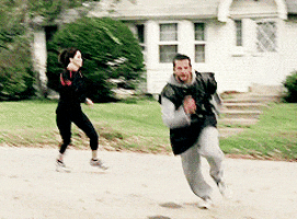 Movie gif. Bradley Cooper as Pat in Silver Linings Playbook wears a gray sweat suit and a black trash bag over his sweatshirt as he runs full speed down the road. Jennifer Lawrence as Tiffany wears a work out outfit as she chases him.