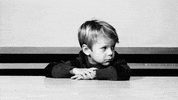 Video gif. A little boy sits with his arms crossed on a table. He turns his head to the side, crinkling his brow in boredom and tapping his fingers impatiently on his arm.