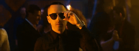 Music video gif. Yandel makes eye contact with a woman walking past the bar and he stares in awe, slowly taking his sunglasses off to get a better look at her.