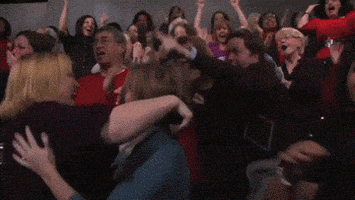 Video gif. A grainy clip shows a crowd going wild with applause as two women in the front jump up and down while hugging each other.