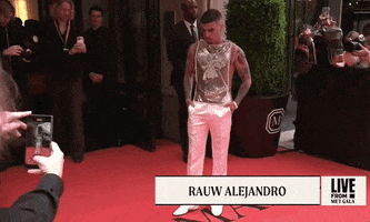Met Gala 2024 gif. Rauw Alejandro wearing a golden sleeveless top and white satin pants, an ensemble by Ludovic de Saint Sernin, turns slowly, adjusting nervously, fully surrounded by cameras.