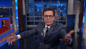 Late Show gif. Stephen Colbert reaches out to us with an open hand on set and says, "Welcome."