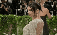 Met Gala 2024 gif. Emily Ratajkowski shows the upper back and front of her long sleeve pale gold Versace gown with a crew neckline. Her hair is swept back into a tight bun with three ends flaring out resembling bird feathers. Two short loose strands hang down and frame her face.