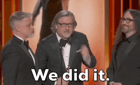Oscars 2024 GIF. Dave Mullins holds up his Oscar on stage and he looks up into the sky and pumps his award up, celebrating his win and thanking the audience. 