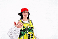 Music Video gif. From Billie Eilish's music video for "LUNCH", Billie is on a stark white background dressed in a white long sleeve shirt and a yellow and black jersey over the top, with a red flat brim hat on. She is pointing at herself while looking into the camera singing her song and getting closer to the lens.