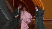 Oscars 2024 gif. Ariana Grande, in an oversized, puffy pink dress, and Cynthia Erivo, in a contrasting forest green dress, run off as Billie Eilish and Finneas O'Connell accept the Best Song award. They awkwardly walk stiffly towards the microphone. 