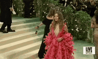Met Gala 2024 gif. We zoom in on Jessica Biel waving and smiling at camera, wearing a hot pink Tamara Ralph Couture full-length gown with a deep-v neckline. The full billowing skirt flares out to a dramatic train. She wears a billowing shrug that matches the pattern of her dress around her back and arms.  