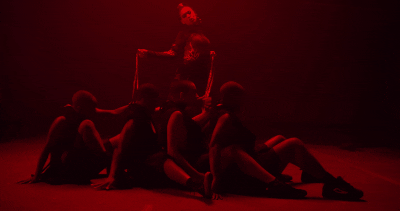 Music video gif. From the video for Bend and Snap, lit by red lights and dressed like a dominatrix, Dizzy Fae holds onto four dancers with leashes as they sit with legs around each other, leaning back.