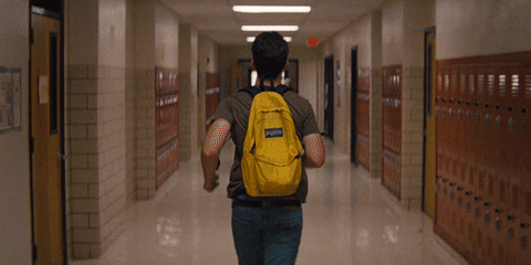 Movie gif. Miles Teller as Sutter Keely in The Spectacular Now runs down a dimly lit, narrow school hallway with a yellow backpack on.