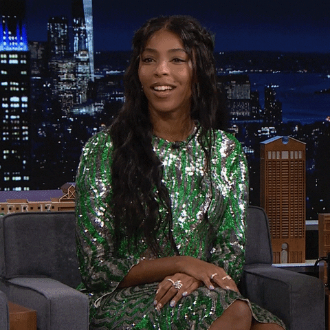 Tonight Show gif. Jessica Williams sits on set with a sparkling silver and green dress. She smiles and waves to the audience, and then blows a series of kisses to them.