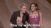 Oscars 2024 GIF. Kate McKinnon and America Ferrera stand on stage and McKinnon is leaned into the microphone, staring intensely without blinking, and says, "You're welcome." 